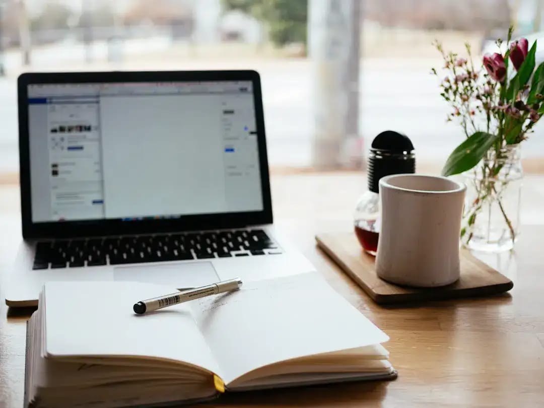 Laptop, notes z piórem i kubek na drewnianym blacie przy oknie