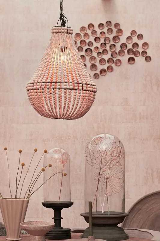 Vor einer rosabraunen Wand hängt ein Leuchter aus Holzperlen, darunter stehen Vasen und Glasglocken mit Trockenblumen.