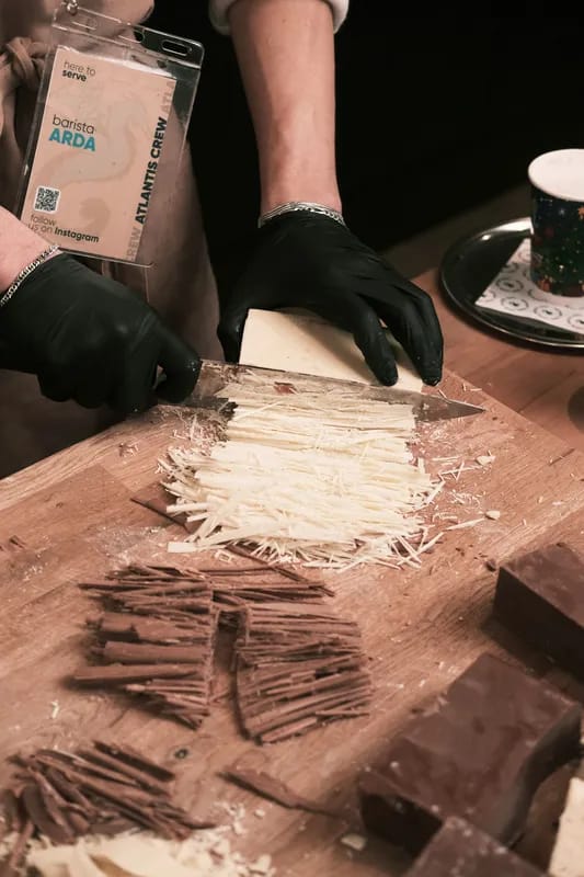 Zwei Hände in schwarzen Handschuhen raspeln an einem Holztisch Schokolade zu feinen Spänen.